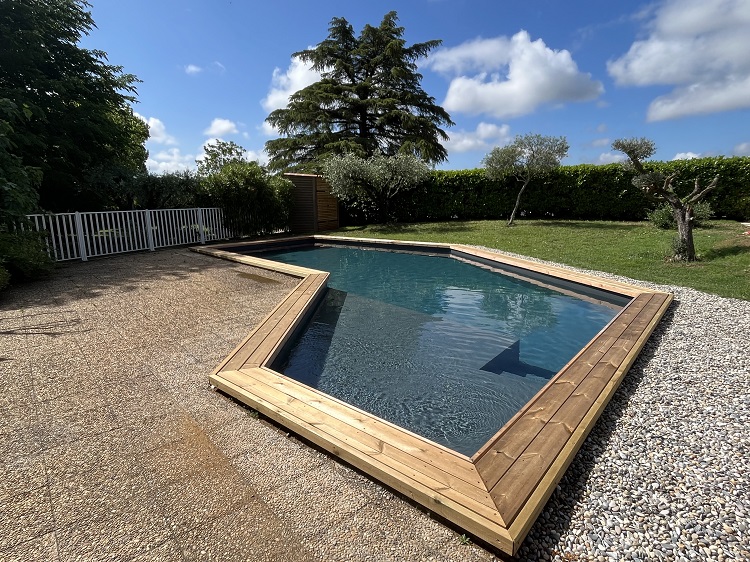 Cette image présente la piscine du Temps des Légendes. Elle est sécurisée par une clôture et arborée par des oliviers et une haie végétale.