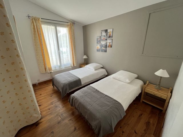 Cette image présente la 2ème chambre de l'étage du Gîte Le Temps des Légendes. Cette chambre lumineuse et spacieuse comporte 2 lit de 90x 200 cm, 2 chevets en pins, et 2 lampes de chevets. Le parquet est en bois massif, et la chambre est dans les tons gris et jaune, et comprend un dressing.