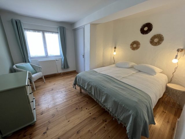 Cette image présente la chambre du rez-de-chaussée du gite Shelby, disposant d'un lit en 160 x 200 cm, de 2 tables de nuit avec 2 lampes de chevet murales de style industiel, une commode, un fauteuil et un placard fermé.