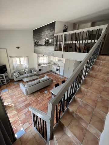 Accès au duplex et sa mezzanine par le séjour Cette image présente le séjour lumineux de 41m2 du gîte Shelby de Le Temps des Légendes, comportant une agréable cuisine équipée et fonctionnelle, un confortable salon télévision avec ses 2 canapés cuirs 3 places, ainsi qu'un espace repas style industriel pour 6 personnes, avec ses escaliers pour accéder à la mezzanine.