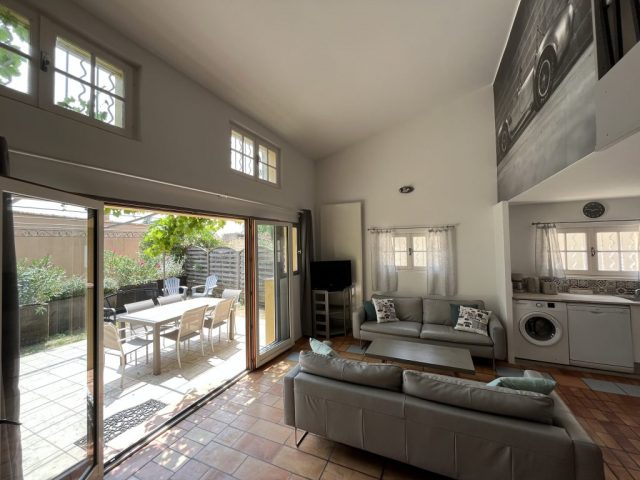 Lumineux séjour ouvert sur la terrasse ombragée et arborée. Cette image présente le séjour lumineux de 41m2 du gîte Shelby de Le Temps des Légendes, comportant une agréable cuisine équipée et fonctionnelle, un confortable salon télévision avec ses 2 canapés cuirs 3 places, ainsi qu'un espace repas style industriel pour 6 personnes.