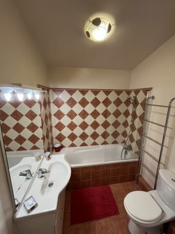Cette image présente la salle de bains comportant sur la gauche un meuble vasque, au fond une baignoire, carrelée dans les tons provençaux beige / terre cuite équipée d'une paroie vitrée et sur la droite les toilettes.