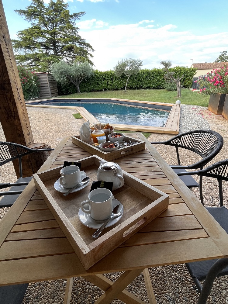 Cette photo présente un plateau petit déjeuner servi sur table du Pool House, face à la piscine et au parc arboré en dernier plan