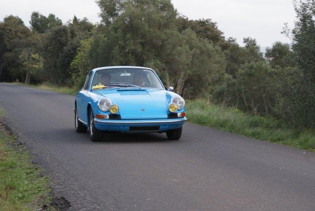 Cette image présente une Porsche 911 bleu sur une route des cévennes avec la garrigues en arrière plan