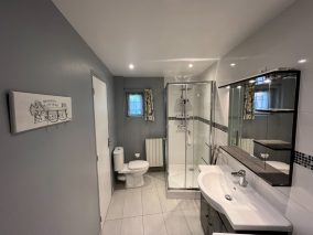 Cette image représente la salle d'eau du gîte Berlinette du Temps des Légendes. Au premier plan, on note le meuble vasque surmonté d'un miroir. en second plan, la grande douche et les toilettes.