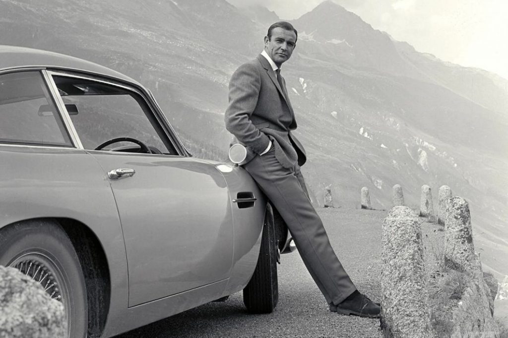 Cette image représente l'agent secret James Bond 007 incarné par Sean Connery, assis sur l'aile de son Aston Martin DB5, extrait du film Goldfinger