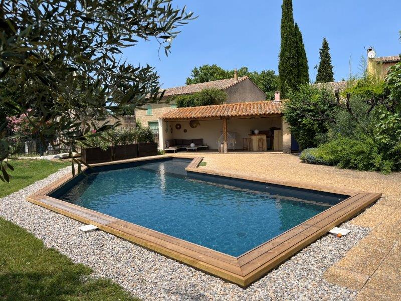 Cette image présente la piscine du Temps des Légendes. Elle est sécurisée par une clôture et arborée par des oliviers et une haie végétale. En arrière plan le Pool house et son salon de détente.